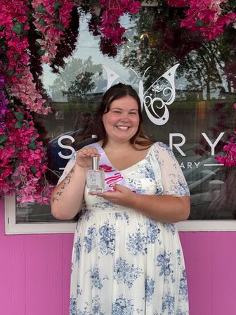 Smiling woman in blue floral dress and sash holding a perfume bottle in front of a pink storefront framed by cascading pink flowers.