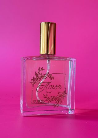 Clear rectangular perfume bottle with shiny gold cap labeled "Amor" and laurel wreath illustration with date 10.18.25, styled on a vibrant hot-pink background — product fragrance photo