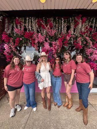 Six women smiling in front of a vibrant pink floral storefront, cowgirl-style outfits with boots and hats, group photo outside a flower-covered shop.