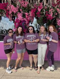Five friends wearing matching 'Nashville' shirts pose holding perfume bottles beneath a vibrant pink and white floral arch outside a pink storefront