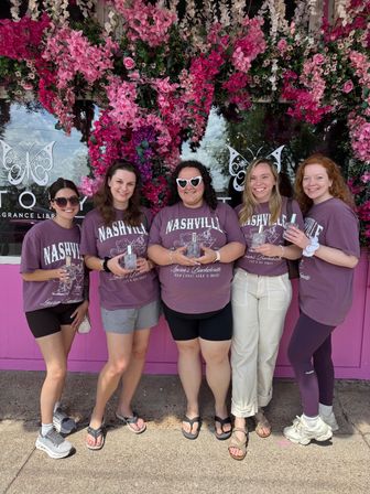 Five friends wearing matching 'Nashville' shirts pose holding perfume bottles beneath a vibrant pink and white floral arch outside a pink storefront