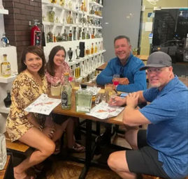 Four friends laughing around a wooden table in an indoor spirits tasting room, sampling colorful cocktails with liquor bottles lined on white shelves behind them.