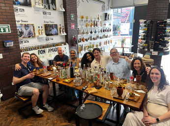 Nine friends smiling and toasting at a tequila and mezcal tasting around a wooden table crowded with bottles, shot glasses and cocktails in a bottle-lined tasting bar.