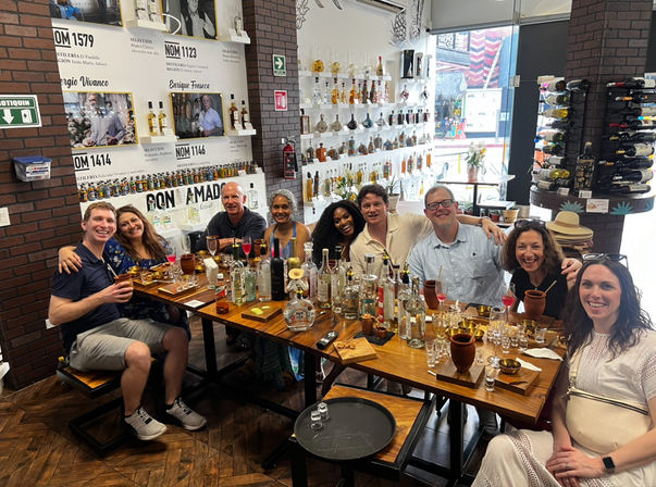Nine friends smiling and toasting at a tequila and mezcal tasting around a wooden table crowded with bottles, shot glasses and cocktails in a bottle-lined tasting bar.
