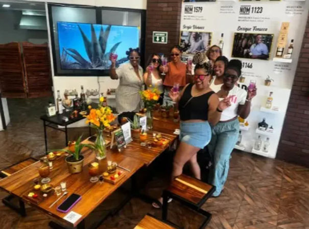 Group of people cheer with pink cocktails in an indoor cocktail-tasting room, gathered around a wooden communal table with flowers, small plates, bottles and an agave-themed display.