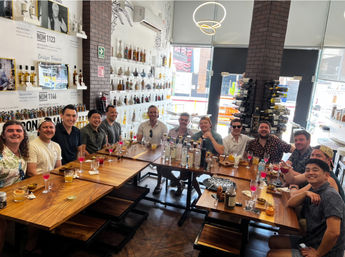 About a dozen people smiling and toasting during a tequila tasting in a bright modern tasting room — long wooden tables filled with bottles, colorful cocktails and tasting glasses, back wall of spirits and a wine rack by large street-facing windows.