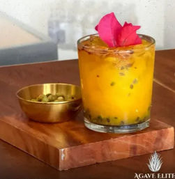 Bright passion-fruit cocktail in a short glass with visible seeds and a pink edible petal garnish, served on a wooden board next to a small brass bowl of cardamom pods — vibrant tropical tabletop styling.