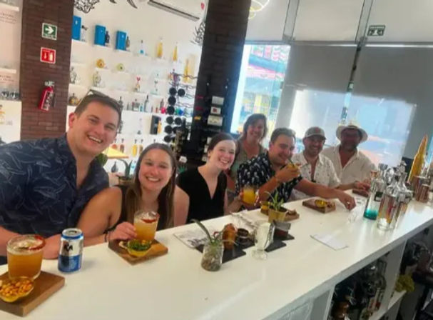 Group of seven friends smiling and enjoying tropical cocktails and small plates while seated along a bright white bar counter in a modern cocktail bar with backlit bottles and daylight windows.