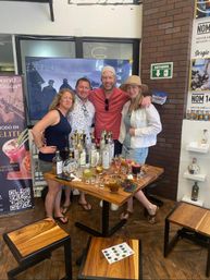 Group of four smiling adults at a mezcal tasting around a wooden table crowded with bottles, shot glasses and colorful cocktails in a cozy Mexican tasting room with brick wall and a 'botiquín' sign