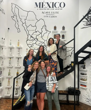 Smiling group of seven women posing on and beside a black metal staircase in a tasting room, in front of a large stylized map of Mexico and shelves of agave spirit bottles — Los Cabos, Mexico.
