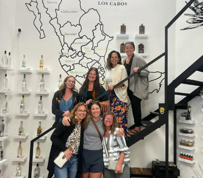 Smiling group of seven women posing on and beside a black metal staircase in a tasting room, in front of a large stylized map of Mexico and shelves of agave spirit bottles — Los Cabos, Mexico.