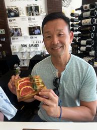 Smiling man in a wine-tasting room holding a wooden board of sliced pink grapefruit and avocado topped with chili powder and rosemary, wine bottles on racks in the background.
