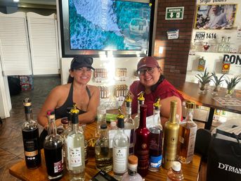 Two smiling women at a wooden tasting table surrounded by assorted tequila and liquor bottles, small agave plants, shelves with product displays and a TV showing ocean waves in a bright tasting room.