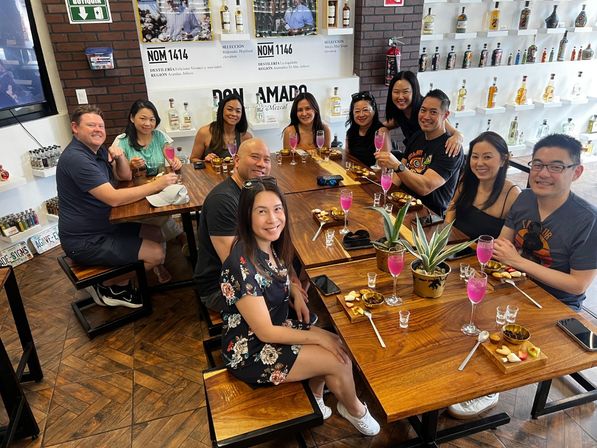 Group of friends at a mezcal and tequila tasting room, smiling around wooden tables with pink cocktails, shot glasses and small snack boards, bottle-lined shelves in background.