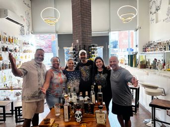 Smiling group of six posing in a bright tequila tasting room and spirits shop, with bottles on shelves, a wooden tasting table in front, modern circular pendant lights and a central brick column.