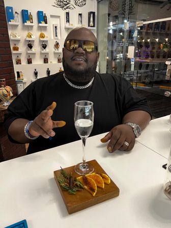 Man in sunglasses and chain at a modern spirits-tasting counter with a champagne flute and a wooden board of charred orange slices, rosemary and spices.