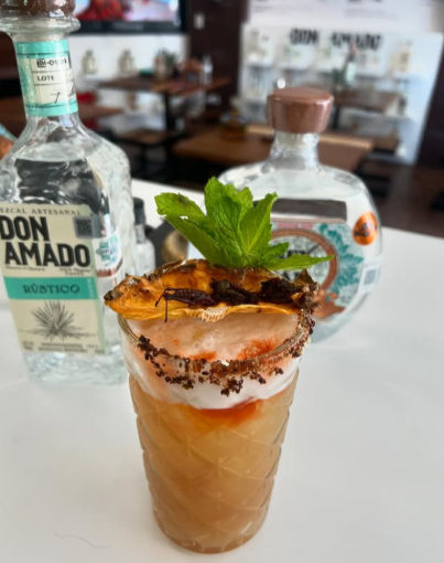 Tall tropical mezcal cocktail in a patterned glass with foamy top, chili-salt rim, dried pineapple and fresh mint garnish on a white bar counter with artisan mezcal bottles blurred in the background.