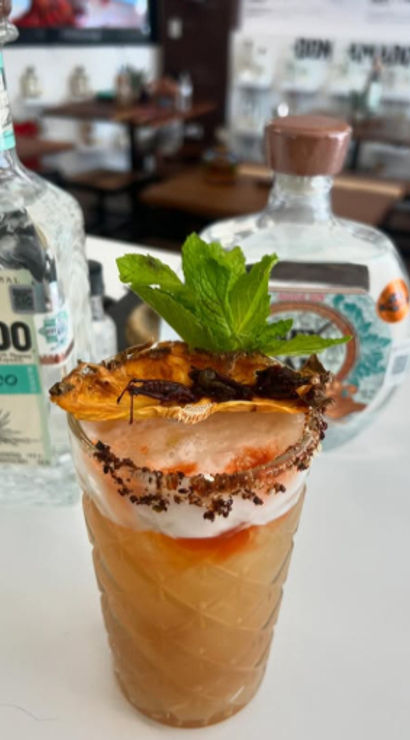 Tall tropical mezcal cocktail in a patterned glass with foamy top, chili-salt rim, dried pineapple and fresh mint garnish on a white bar counter with artisan mezcal bottles blurred in the background.