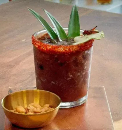 Mexican-style spicy tomato cocktail (michelada) in a chili-rimmed glass, topped with pineapple leaves and a lime wedge, served with a small bowl of peanuts on a wooden table.