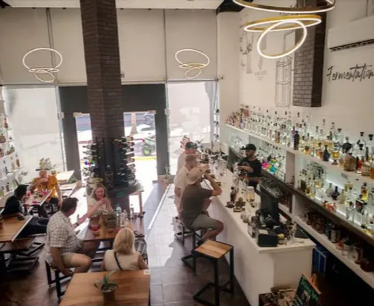 Sunlit modern cafe-bar interior with high ceilings and circular pendant lights, long backlit shelves of liquor bottles, patrons seated at wooden tables and a busy bar by large front windows