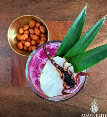 Top-down view of a vibrant magenta tropical cocktail on a wooden board, garnished with pineapple leaves, a pear slice and a whole scorpion, beside a small brass bowl of spiced peanuts.
