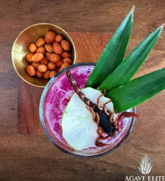 Top-down view of a vibrant magenta tropical cocktail on a wooden board, garnished with pineapple leaves, a pear slice and a whole scorpion, beside a small brass bowl of spiced peanuts.