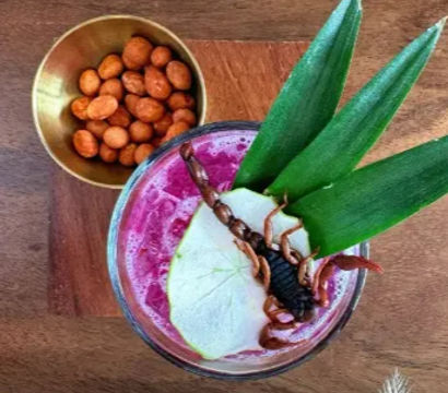 Top-down view of a vibrant magenta tropical cocktail on a wooden board, garnished with pineapple leaves, a pear slice and a whole scorpion, beside a small brass bowl of spiced peanuts.