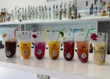 Row of colorful tropical cocktails and fruit smoothies served in clear pouch cups with straws and small floral garnishes lined up on a white bar counter, blurred bottles on shelves in the background