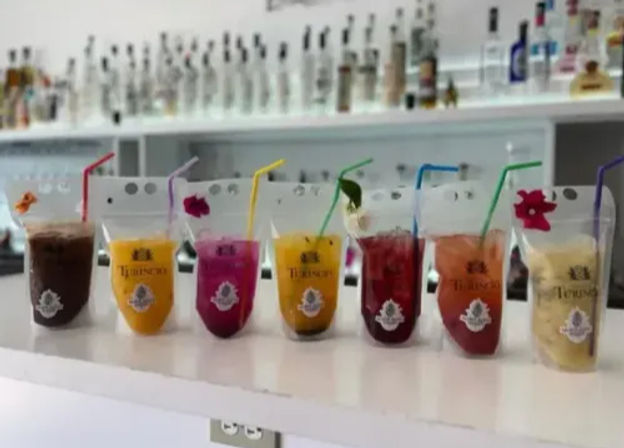 Row of colorful tropical cocktails and fruit smoothies served in clear pouch cups with straws and small floral garnishes lined up on a white bar counter, blurred bottles on shelves in the background