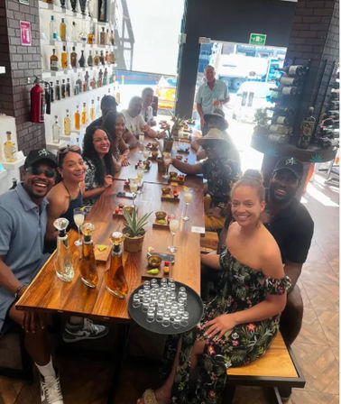 Smiling group of friends at a sunlit indoor bar enjoying a tequila tasting around a long wooden communal table with shot glasses, cocktails, agave decor and shelves of liquor bottles