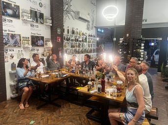 Group of adults seated at long wooden tables in a lively tequila tasting room, raising bottles topped with sparklers amid shelves of spirits and wine racks under modern lighting