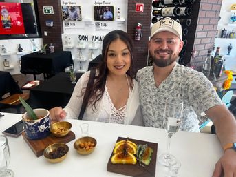 Smiling couple enjoying a mezcal tasting in a bright tasting room with bottles on display, brass snack bowls, a citrus garnish board, and a tall tasting glass on the bar.