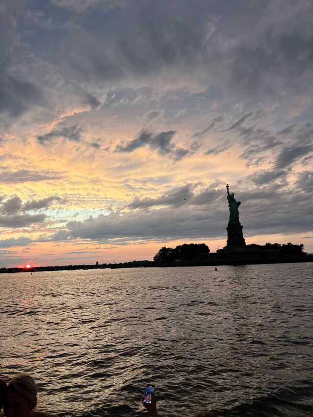 Statue of Liberty silhouette in New York Harbor at sunset with dramatic clouds, rippling water, and a passenger holding up a phone in the foreground