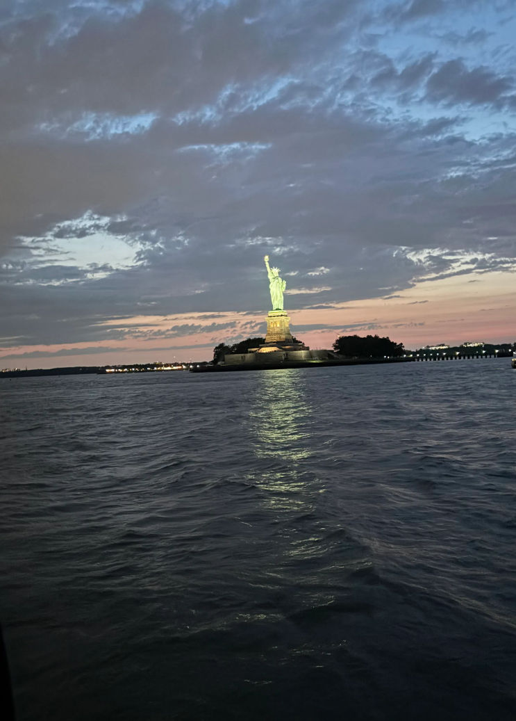 Statue of Liberty illuminated at dusk, glowing green atop its pedestal in New York Harbor with rippling water and a colorful sunset sky.