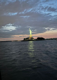 Statue of Liberty illuminated at dusk, glowing green atop its pedestal in New York Harbor with rippling water and a colorful sunset sky.