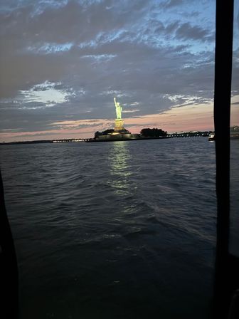 Dusk view of the illuminated Statue of Liberty on Liberty Island, glowing green against a pink-blue sunset sky with its light reflecting on New York Harbor waters from a nearby boat.