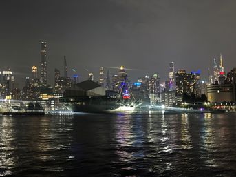 Nighttime waterfront with an illuminated museum ship in the foreground and the glowing New York City skyline reflecting colorful lights on rippling water.