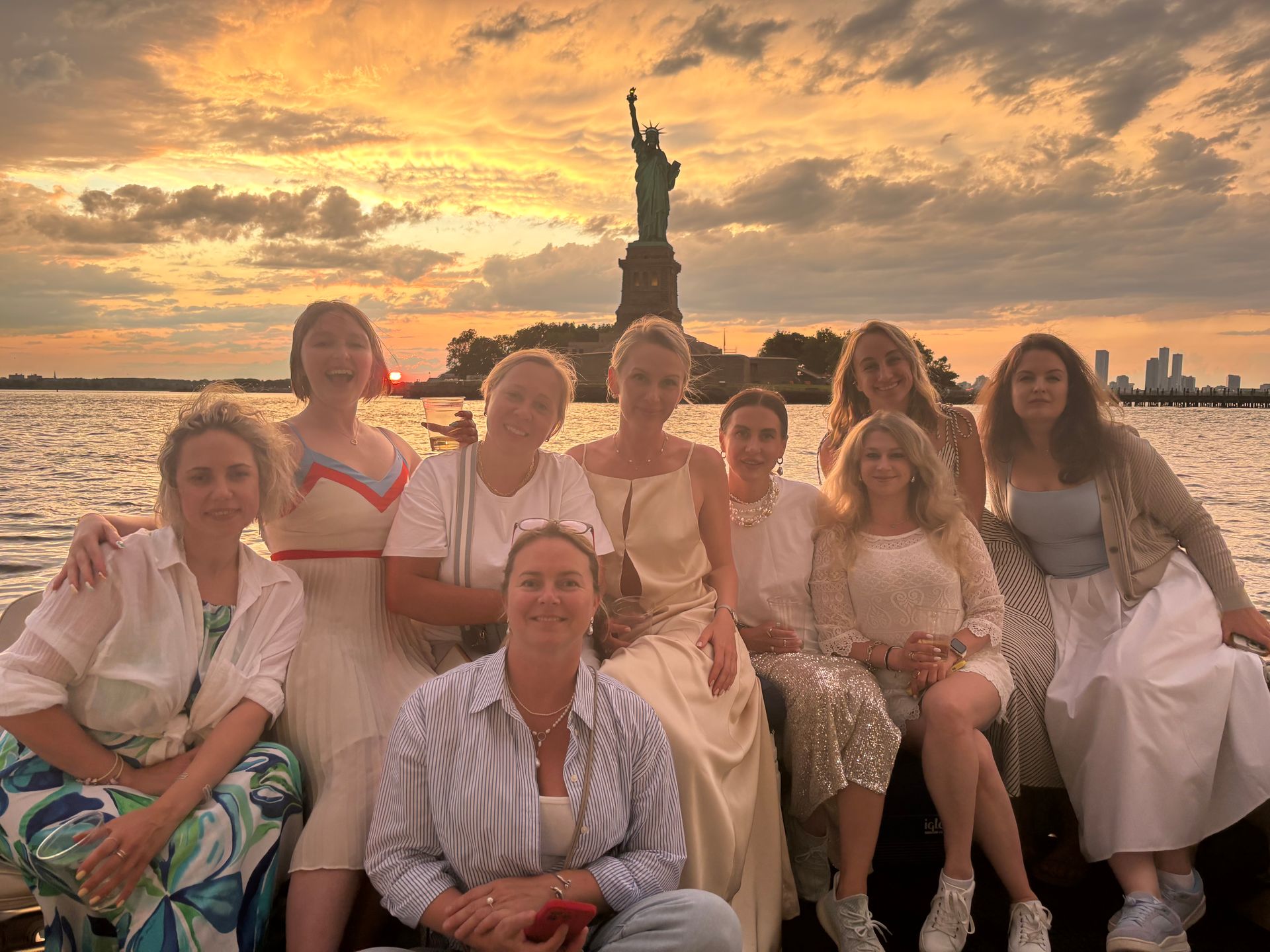 Group of women posing on a boat in New York Harbor at sunset with the Statue of Liberty silhouetted against a golden sky and the Manhattan skyline in the distance.