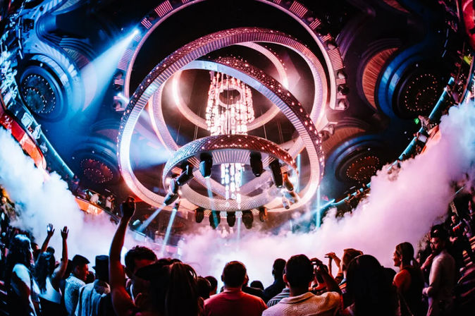 Packed nightclub dance floor beneath a spiral LED chandelier with colorful beams, fog machines, and an energetic nightlife crowd