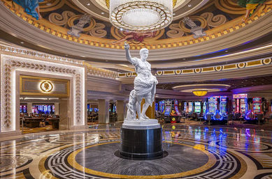 Luxurious casino lobby with a white Roman-style statue beneath a crystal chandelier, ornate painted dome, polished marble floor and colorful slot machines.