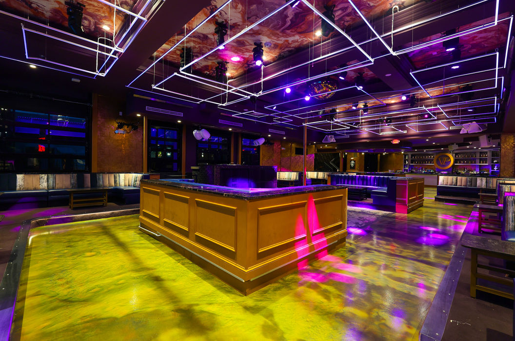 Modern nightclub interior with glowing yellow-green dance floor, central wooden bar, purple neon grid ceiling lights, and plush lounge seating