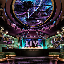 Upscale nightclub interior with swirling neon LED ceiling, illuminated stage with large glowing letters, curved VIP booths and colorful light beams over a dark dance floor.