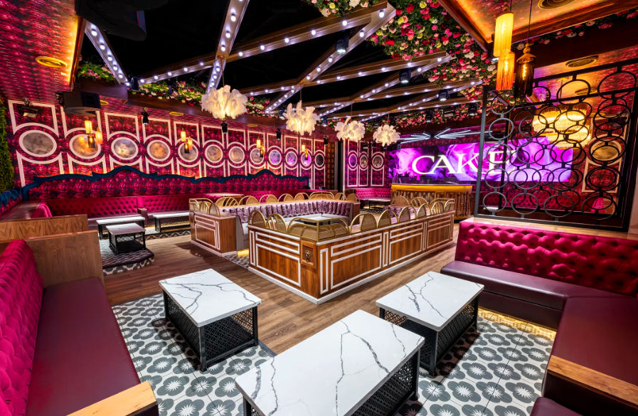 Vibrant luxury nightclub lounge interior with tufted velvet banquettes, marble-top cocktail tables, central VIP booth, floral ceiling accents, exposed-beam LED lighting and colorful backlit bar — stylish urban nightlife setting.