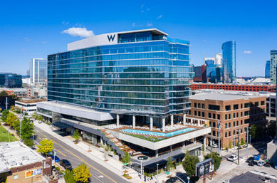 Gleaming glass high-rise hotel with rooftop pool and sun loungers overlooking a vibrant downtown skyline and tree-lined street under a bright blue sky