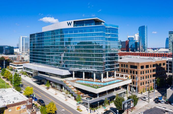 Gleaming glass high-rise hotel with rooftop pool and sun loungers overlooking a vibrant downtown skyline and tree-lined street under a bright blue sky