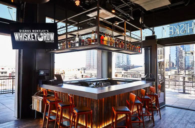 Sunlit rooftop whiskey bar with a square central counter, suspended bottle racks, red bar stools and a downtown skyline view.