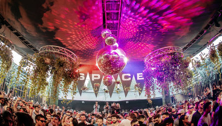 Packed outdoor covered terrace dance party with oversized mirrored disco balls casting pink and purple lights, hanging greenery and a lively crowd enjoying nightlife vibes.