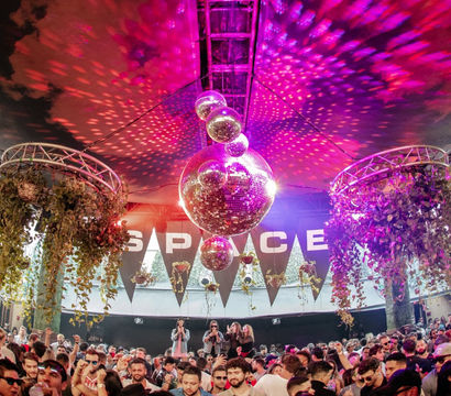 Packed outdoor covered terrace dance party with oversized mirrored disco balls casting pink and purple lights, hanging greenery and a lively crowd enjoying nightlife vibes.