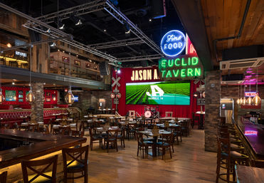 Industrial-chic downtown dining and live music venue interior with neon signs, stage with drum kit, red curtain, wooden floors, scattered tables and a long bar.