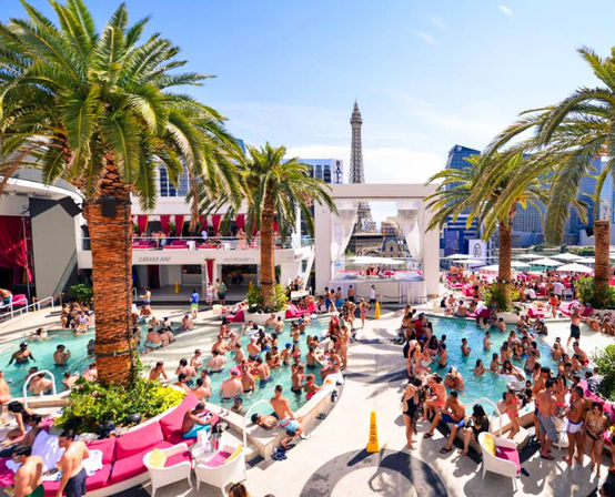 Vibrant daytime Las Vegas Strip pool party with crowded resort pools, palm trees, pink cabanas and lounge seating, framed by the Eiffel Tower replica and city skyline in the background